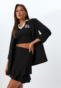 Zwarte cropped top met witte kraag en grafische print, gecombineerd met een zwarte gelaagde rok. De outfit wordt aangevuld met een zwart blazer.
