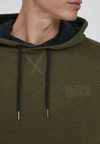 Olivgrüner Kapuzenpullover mit navyblauer Innenseite, schwarzen Kordeln und geprägt Logo-Detail auf der Brust. Strukturierter Stoff.