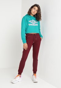 Teal cropped hette med hvit logo, kombinert med burgunder joggebukser med hvite sidestriper. Modellen har på seg hvite sneakers med fargerike detaljer.