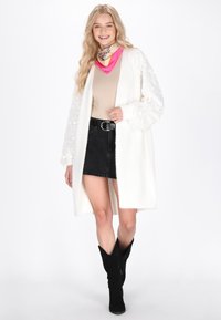 Bela cardigan z teksturiranimi rokavi, bež top in črno džins krilo. Dodatki vključujejo roza in bež šal ter črne zaščitne čevlje iz semiša.