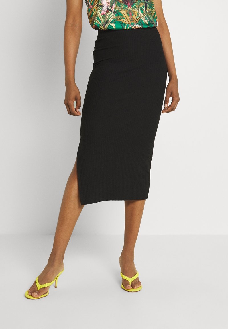 Jupe midi noire côtelée avec une taille haute et une fente latérale, associée à des talons jaunes à lanières. Présente une texture lisse et un design ajusté.