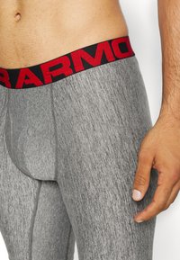 Graue sportliche Leggings mit strukturierter Oberfläche und einem schwarzen Bündchen mit roter Schrift. Nahaufnahme, die den Taillenbereich und den Stoff hervorhebt.