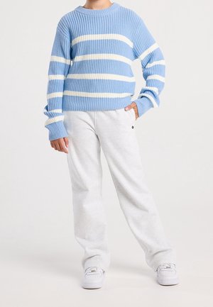 Personne portant un pull en tricot bleu clair à rayures horizontales blanches, un pantalon blanc ample et des baskets blanches, debout avec une main dans la poche.