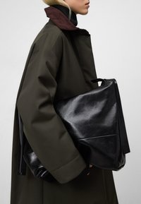 Borsa in pelle nera con finitura lucida tenuta sotto un cappotto verde con collo in velluto a coste, che mostra un design strutturato e uno stile minimalista.