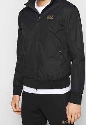 Hombre con chaqueta negra con cremallera EA7 Emporio Armani con puños elásticos y pantalones negros a juego con parche de logo.