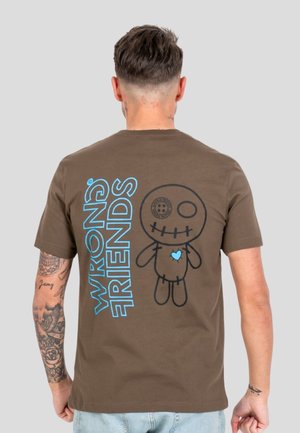Mężczyzna zwrócony tyłem, ubrany w brązowy T-shirt z niebieskim napisem "WRONG FRIENDS" oraz kreskówkową, zszywaną lalką z guzikiem zamiast oka i niebieskim sercem.