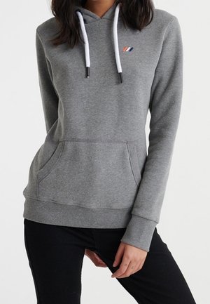Femme portant un sweat à capuche gris ajusté avec des cordons blancs et un petit logo tricolore sur la poitrine, associé à un pantalon noir.