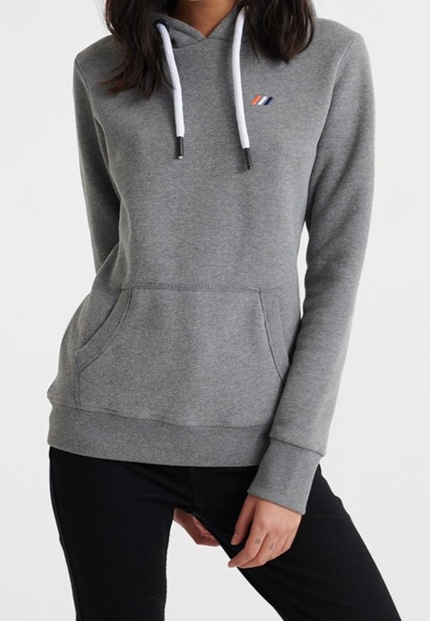 Femme portant un sweat à capuche gris ajusté avec des cordons blancs et un petit logo tricolore sur la poitrine, associé à un pantalon noir.