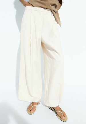 Personne portant un pantalon blanc ample et des sandales beige type tongs, debout devant un fond clair uni.