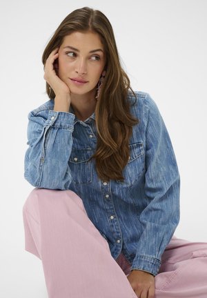 Kobieta z długimi brązowymi włosami, ubrana w niebieską jeansową kurtkę i różowe spodnie, opierająca twarz na dłoni i patrząca na bok.