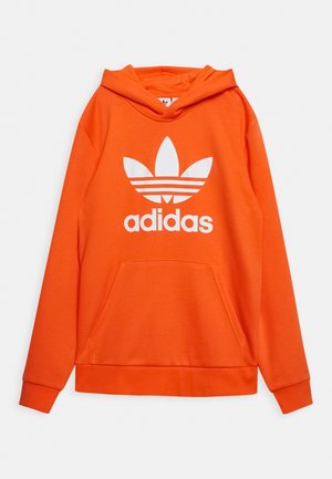 Sweat à capuche orange vif avec poche kangourou avant et grand logo Adidas trèfle blanc ainsi que texte sur la poitrine.