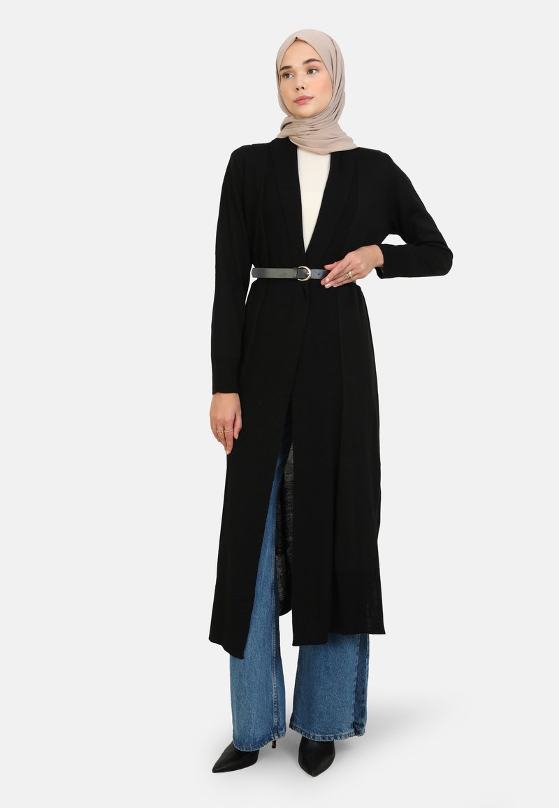 Zwarte lange cardigan, gebreid materiaal, open voorkant met een ceintuur in de taille, gecombineerd met een lichtgekleurde top en wijde denim jeans.