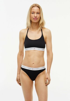 Frau steht und trägt schwarzen Tommy Hilfiger Bralette und Slip mit weißen elastischen Bändern mit Markenname und Logo.