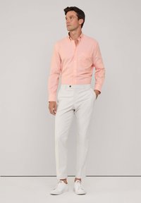 Camisa de botones rosa claro con un bolsillo en el pecho, combinada con pantalones ajustados blancos y zapatillas blancas, frente a un fondo neutro.