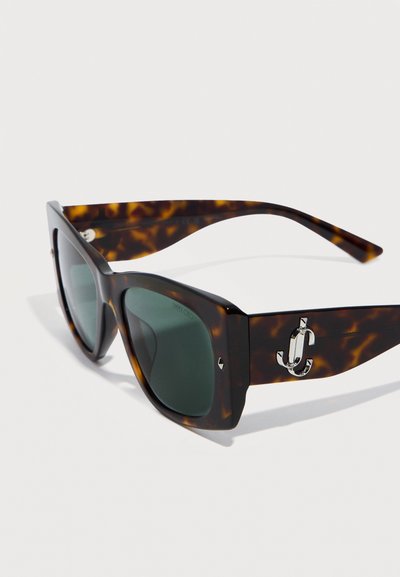 Ορθογώνια γυαλιά ηλίου με καφέ σκελετό και σκουρόχρωμους φακούς, με ασημένιο λογότυπο "JC" στα πλατιά πλαϊνά τους, παρουσιάζονται σε λευκό φόντο.