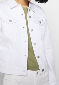 Valkoinen farkkutakki hopea-napeilla, puettuna oliivinvihreän paidan päälle, yhdistetty valkoisiin farkkuihin, kuva kaulasta lantioon.