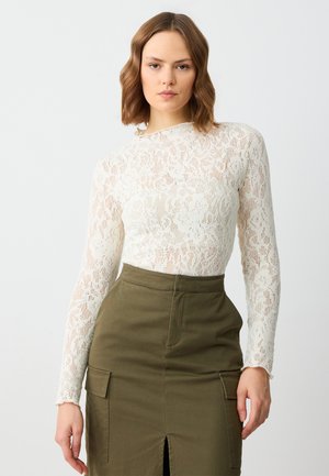 Haut à manches longues en dentelle couleur ivoire avec un motif floral, associé à une jupe verte olive taille haute avec une fente avant et des poches latérales.