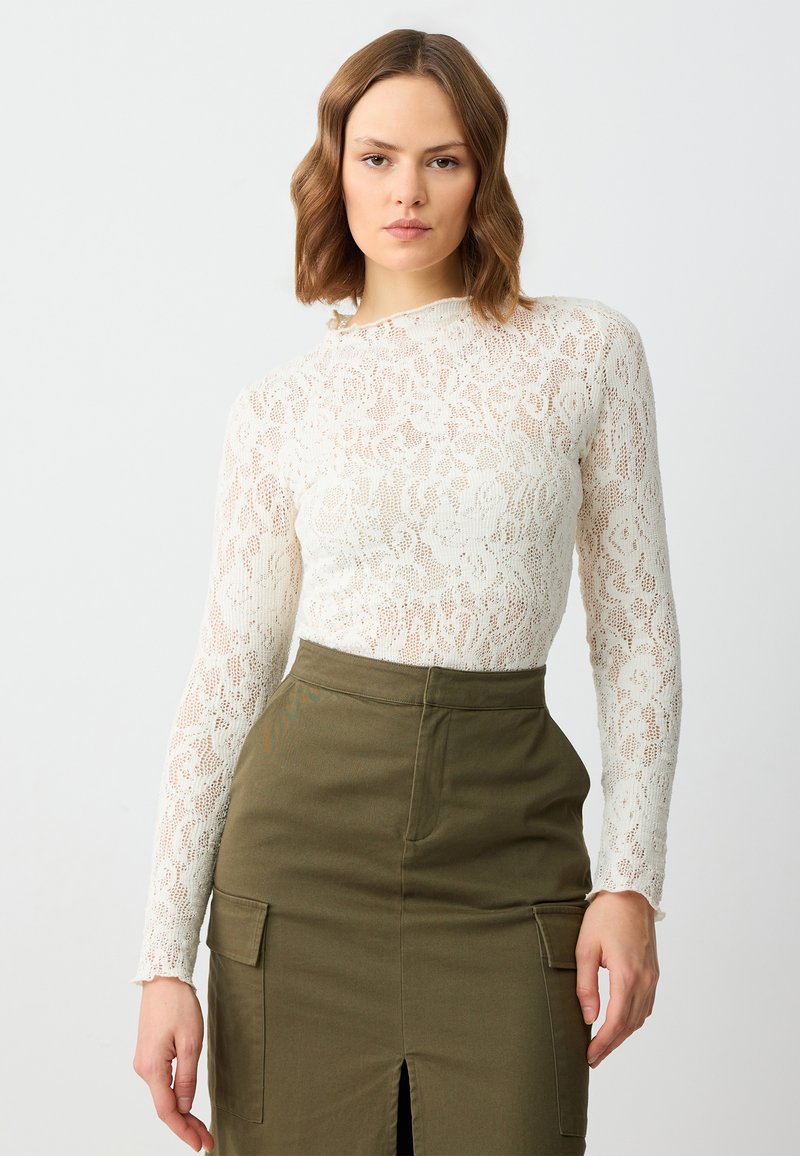 Haut à manches longues en dentelle couleur ivoire avec un motif floral, associé à une jupe verte olive taille haute avec une fente avant et des poches latérales.