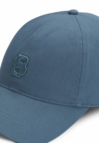 Blue cap made of structured fabric, featuring a curved brim and embroidered logo on the front. Textured ribbed design and tonal stitching.
