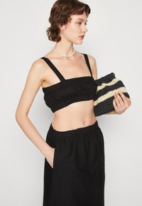 Musta lyhyt toppi leveillä olkaimilla ja ryppytetyllä kankaalla; yhdistettynä mustiin hameisiin. Pitelee raidallista neulottua clutch-laukkua mustana ja kermanvalkoisena.