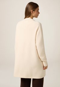 Długi, kremowy kardigan o luźnym kroju, z prążkowanymi mankietami i gładką fakturą, sięgający za biodra. Prosty, minimalistyczny design.