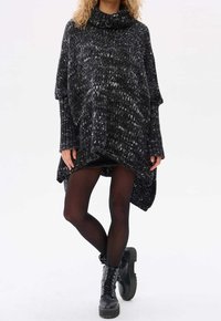 Schwarz-weiß gepunkteter Strick-Poncho mit hohem Kragen und übergroßen Ärmeln, kombiniert mit schwarzen Strumpfhosen und Schnürstiefeln.