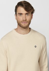 Beige geribbelde gebreide trui met een ronde halslijn en een klein zwart logo op de linkerkant van de borst. Zachte textuur, casual pasvorm en lange mouwen.