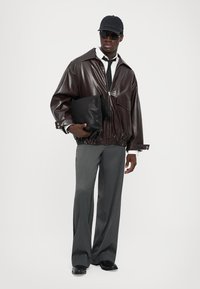 Giacca in finta pelle marrone con una vestibilità rilassata, pantaloni a pieghe grigi, cappellino nero e occhiali da sole, mentre tiene una borsa clutch nera in morbido materiale.