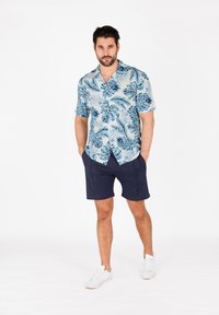 Korte mouwen, lichtblauw Hawaiiaans shirt met groene bladeren, gecombineerd met donkerblauwe shorts en witte sneakers. Eenvoudig, casual ontwerp.