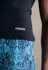 Čierny atletický top s nápisom "STRONGER"; skombinovaný s modrými legínami s vzorom pripomínajúcim hada a textúrovanou tkaninou.