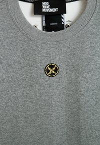 Sudadera gris de tela de mezcla de algodón con cuello redondo, que presenta un parche bordado circular en negro y dorado en el pecho.
