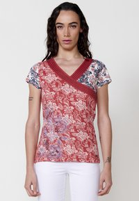 Rotes kurzärmliges Oberteil mit V-Ausschnitt, verziert mit floralen Mustern in Blau und Weiß. Strukturierter Überwurf-Design mit kontrastierenden Ärmeln.