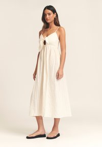 Valge maksikleit, mille ees on lips, õhukesed rihmad ja tekstureeritud kangas; kombineeritud mustade lameda tallaga sandaalidega.