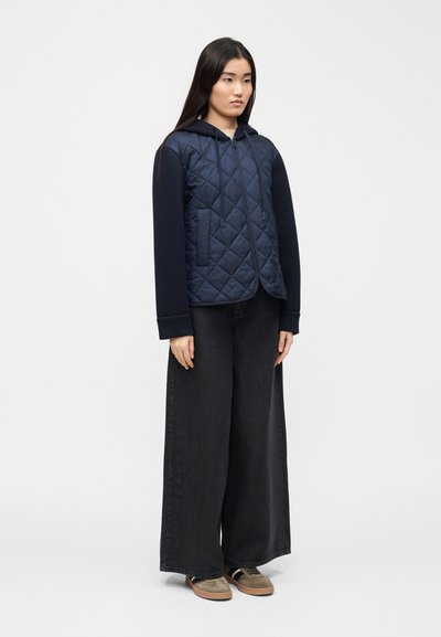 Femme portant un sweat à capuche matelassé bleu marine avec fermeture éclair, pantalon large noir et baskets beige, debout devant un fond blanc uni.