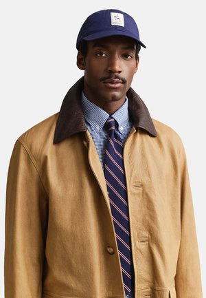 Man draagt een beige leren jas met een donkere kraag, een blauw geruit overhemd, een gestreepte stropdas en een marineblauwe pet met een geborduurd logo.