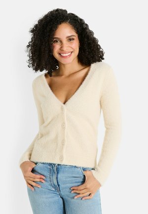 Cardigan de color crema, esponjoso, con un escote en V profundo, seis botones frontales y mangas largas, combinado con jeans de mezclilla. Textura suave y ajuste relajado.