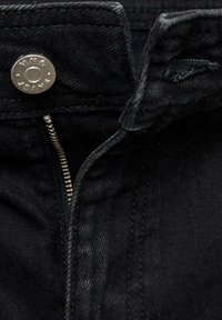 Jean en denim noir en gros plan montrant une fermeture éclair en métal partiellement dézippée et un bouton en métal marqué « VNG JEANS » sur la ceinture.