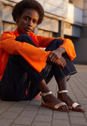 Jong persoon met kort krullend haar, gekleed in een oranje top met lange mouwen, donkere spijkerbroek en metalen sandalen, zittend op een verharde buitenoppervlakte.