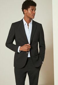 Veste de costume noire avec des revers crantés, fermeture à un bouton et deux poches avant, associée à une chemise rayée bleu clair.