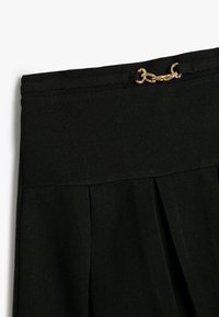 Jupe en tissu plissé noir avec une texture lisse. Elle présente un accent en chaîne dorée au niveau de la taille pour un maintien sécurisé.