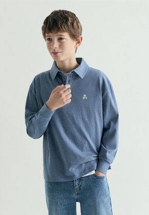 Dreng med kort brunt hår iført en blå polo med lange ærmer med et kranie-logo og blå jeans, stående mod en ensfarvet baggrund.