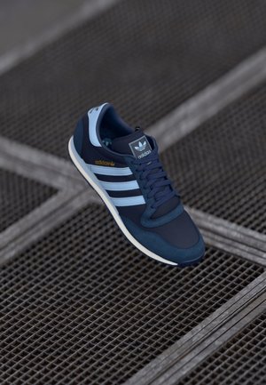 Basket Adidas bleu et marine avec des bandes blanches et un logo, placée sur un fond de grille métallique sombre.