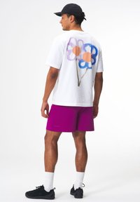 Weißes T-Shirt mit kurzen Ärmeln, das ein buntes Blumenmuster auf dem Rücken zeigt, kombiniert mit leuchtend lila Shorts und schwarzen Sportschuhen.