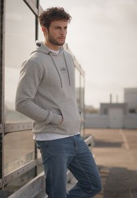 Lichtgrijze hoodie met een voorzak, koordjes en bedrukte logo. Gecombineerd met blauwe denim jeans, staand tegen een glazen achtergrond.