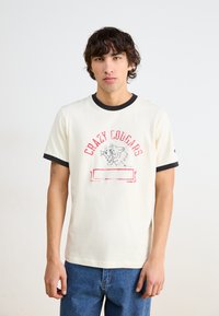 Tricou cu mâneci scurte, de culoare albă-crem, cu guler și manșete negre, având un imprimeu grafic roșu cu un cougar și textul "Crazy Cougars."