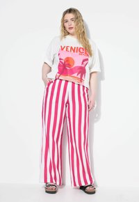 Kvinna i vit oversized t-shirt med "Venice Beach" grafik och rosa och vita vertikalt randiga vida byxor, stående mot vit vägg.