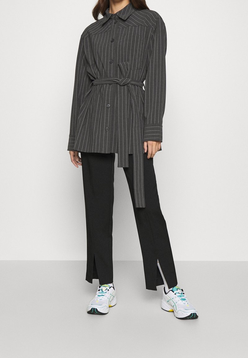 Chaqueta de camisa a rayas negras y grises con cintura ajustable, combinada con pantalones negros de frente partido y zapatillas blancas con acentos de colores.