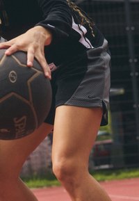 Pantalones cortos atléticos en negro y gris con textura de malla, con un logo de Nike. Una mano sostiene un balón de baloncesto redondo, parcialmente visible en primer plano.