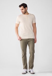 Beige kortermet henley-skjorte med tre knapper, kombinert med olivengrønne bukser og hvite sneakers med svarte striper. Glatt bomullstekstur.
