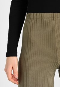 Khaki prążkowane legginsy z elastycznym paskiem w talii, z pionowymi paskami i gładką teksturą. Ręka oparta na tkaninie.